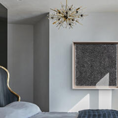 A white bedroom with a decorative chandelier and a velvet grey bed with gold trimmings
